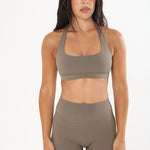 A woman modeling a matching two-piece activewear set in a solid taupe color, featuring a scoop-neck sports bra and high-waisted leggings, against a plain white studio background.