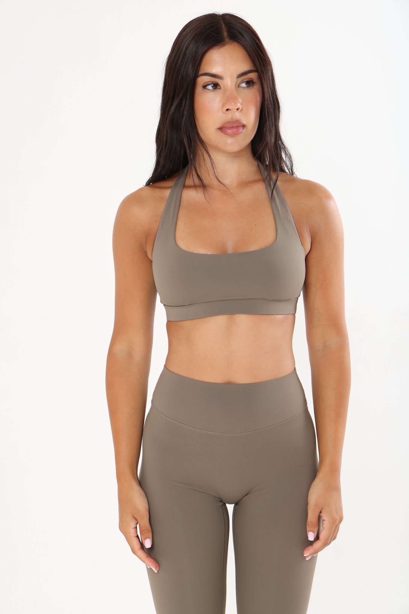 A woman modeling a matching two-piece activewear set in a solid taupe color, featuring a scoop-neck sports bra and high-waisted leggings, against a plain white studio background.
