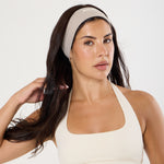 A woman modeling a beige athletic headband and matching beige sports bra against a plain white studio background.