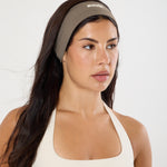 A woman wearing a taupe athletic headband with a small white logo and a cream sports bra, photographed against a clean white studio background.