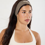 A woman wearing a taupe athletic headband with a small white logo and a cream sports bra, photographed against a clean white studio background.