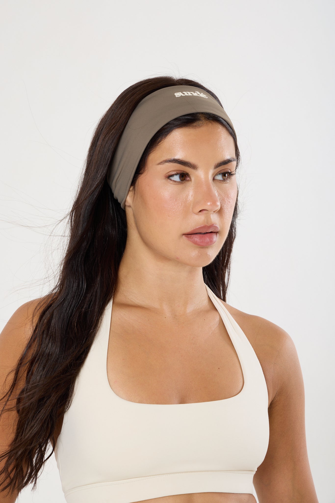 A woman wearing a taupe athletic headband with a small white logo and a cream sports bra, photographed against a clean white studio background.