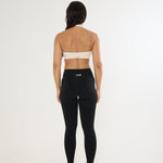 A woman is shown from behind wearing black high-waisted leggings with a small white logo on the back waistband, paired with a beige sports bra and white sneakers, against a plain white studio background.