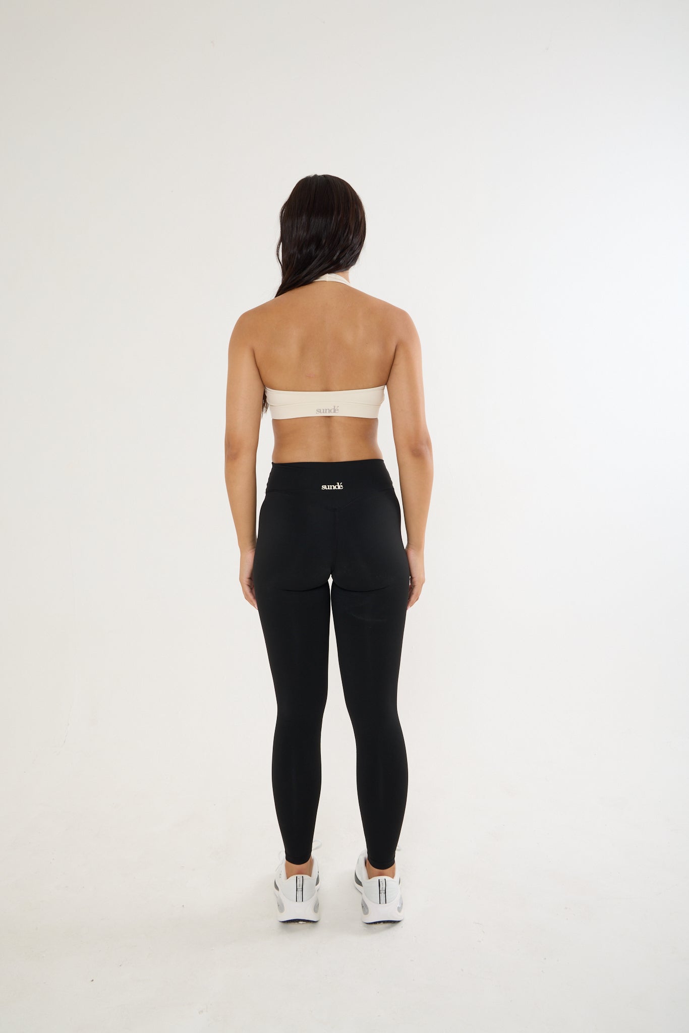 A woman is shown from behind wearing black high-waisted leggings with a small white logo on the back waistband, paired with a beige sports bra and white sneakers, against a plain white studio background.